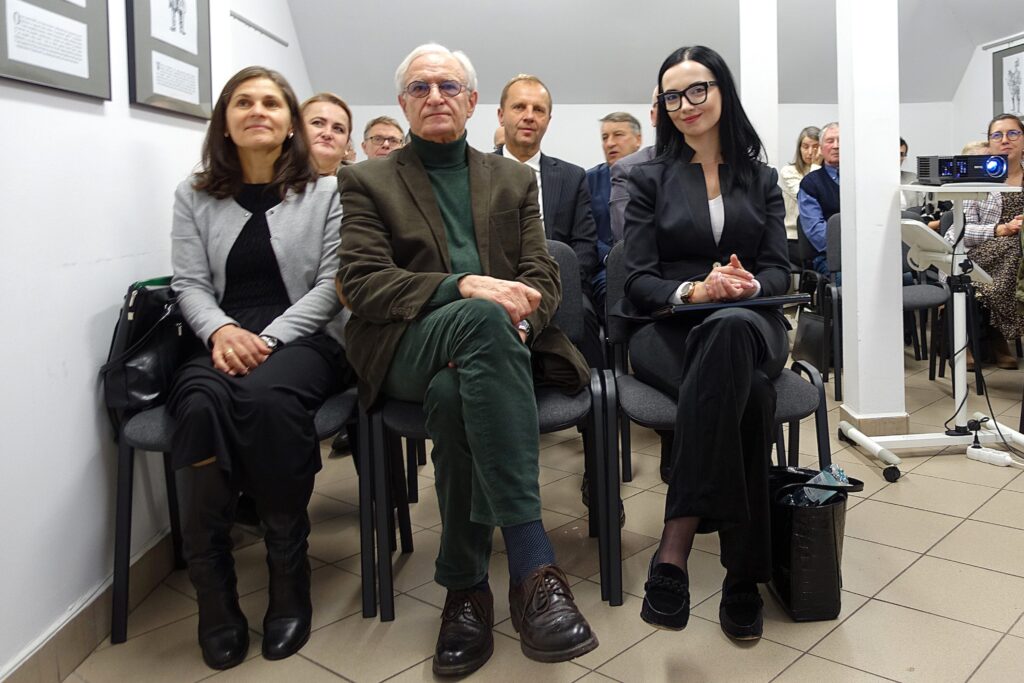 Grupa ludzi siedzących na szarych krzesłach w jasnym pomieszczeniu, z przodu mężczyzna w zielonym garniturze i dwie kobiety, jedna w jasnoszarej marynarce, druga w czarnym garniturze i okularach.