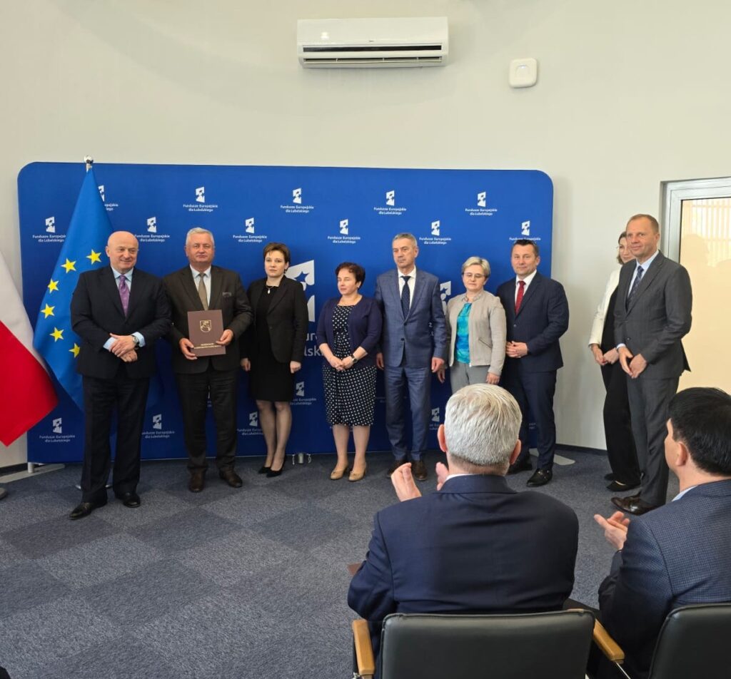 Osiem osób w formalnych strojach stoi na tle niebieskiego banera z białym napisem, obok dwie flagi. Dwie osoby siedzą przodem do grupy i klaszczą.