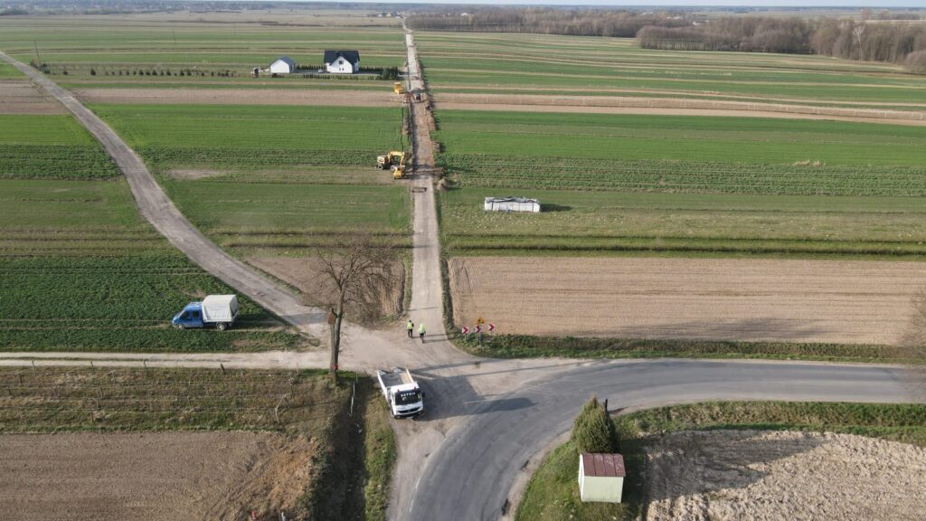 Droga gruntowa przecinająca zielone i brązowe pola, ciężarówka i niebieski samochód, dwa stojące drzewa, dom i maszyny budowlane na tle pól.