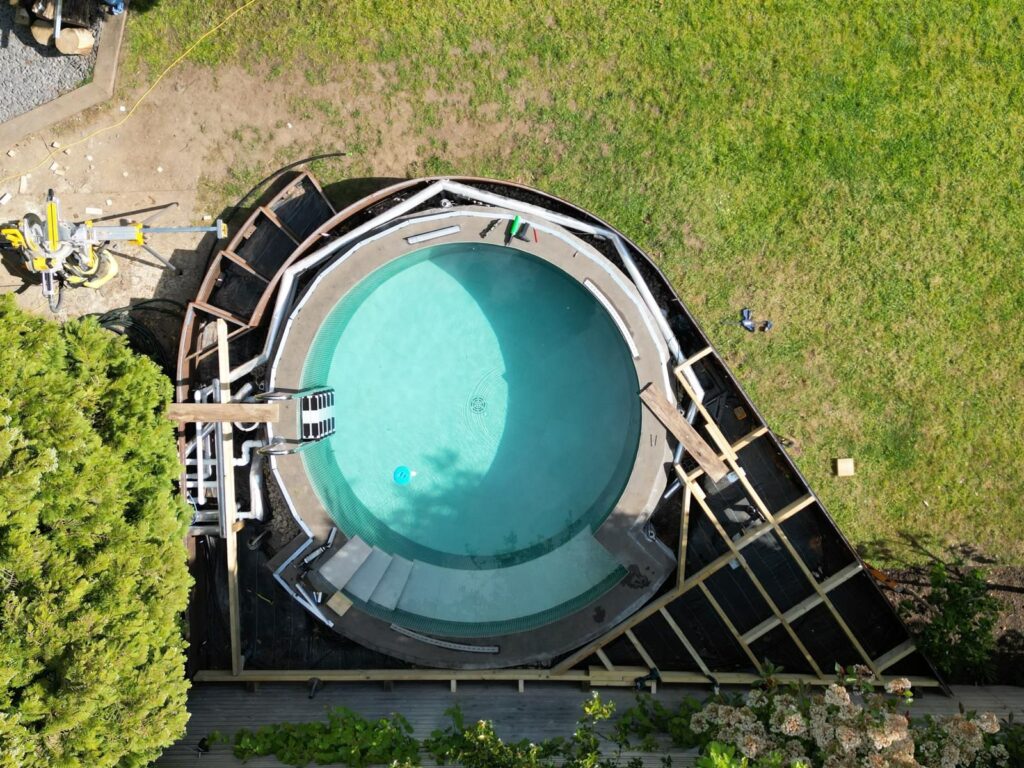 Basen ogrodowy z niebieską wodą, częściowo zacieniony, otoczony konstrukcją drewnianą i trawnikiem.