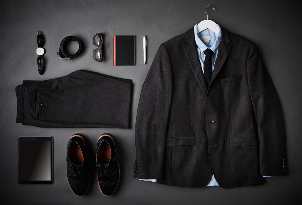 Czarny garnitur z niebieską koszulą i czarnym krawatem na wieszaku, czarne spodnie, buty, zegarek, pasek, okulary, notes, długopis, tablet na ciemnym tle.