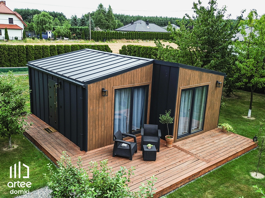 Nowoczesny domek z ciemnoszarym dachem i drewnianymi panelami, taras z dwoma czarnymi fotelami i stolikiem, otoczony ziele...